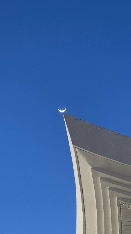 The tip of a white roof against a deep blue sky with a thin crescent moon