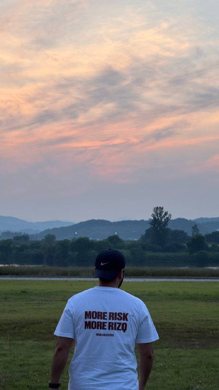 A man in a white T-shirt seen from behind, looking across a field at dusk