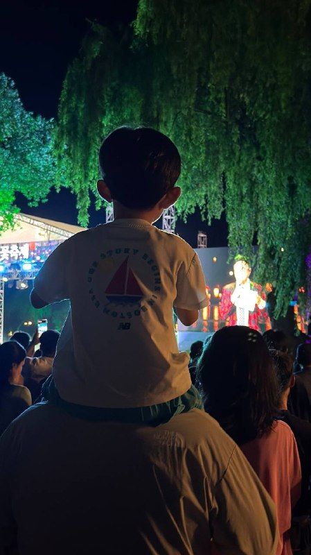 A boy on his father's shoulders watching a lit stage under willow trees at night