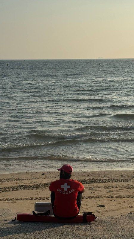 A lifeguard in red sitting on the sand, facing a hazy pastel sea