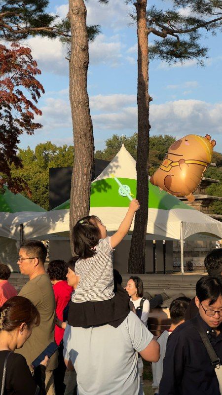 A child on a parent's shoulders reaching towards a giant balloon among pines